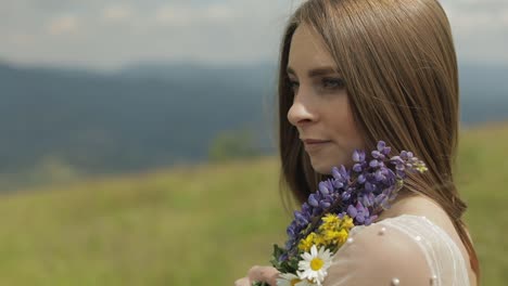 woman in a white dress holding a bouquet of flowers stands in a field with mountains in the background