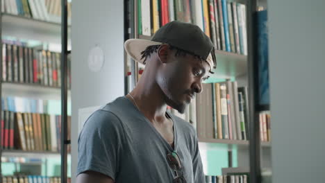 Library attendant wearing cap and casual t shirt standing close to shelf, looking focused in brightly lit library interior with rows of books, creating scene of concentration