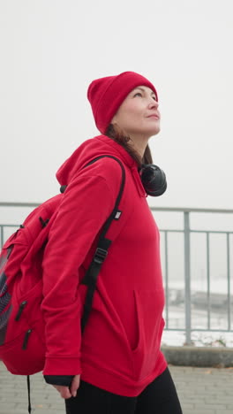 side view of athlete walking near iron railing wearing red beanie and jacket with headphones and backpack, snowy urban scenery with bridge and blurred buildings in foggy winter atmosphere
