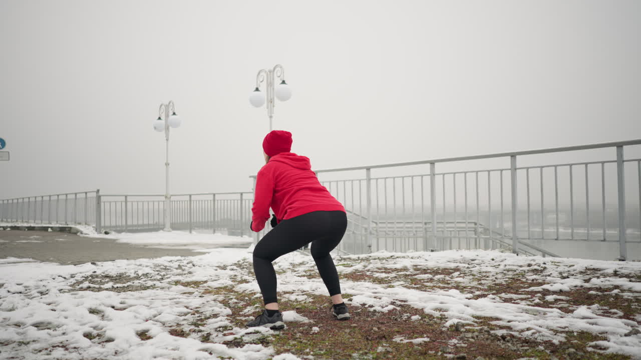 은 후디와 검은 장갑을 입고 눈 인 지상에서 스 점프를 수행하는 코치가 겨울에 야외에서 운동합니다. 장면은 얼어붙은 분위기, 먼 기념비, 그리고 눈 은 언덕의 나무가 특징입니다.
