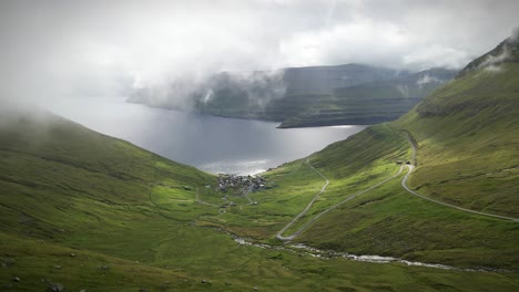 una pintoresca vista de funnings, las islas feroe, con un pequeño pueblo enclavado en un valle verde, rodeado de carreteras sinuosas, montañas escarpadas y una serena ensenada del océano bajo un cielo temperamental