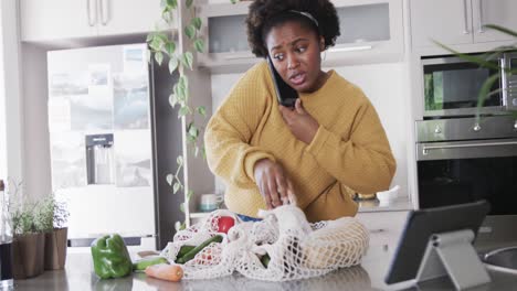 Feliz-Mujer-Afroamericana-Hablando-Por-Teléfono-Inteligente-Desempacando-Comestibles-En-La-Cocina,-Cámara-Lenta