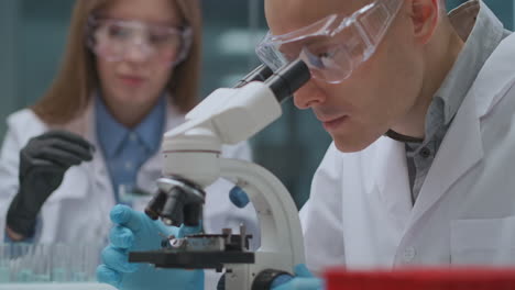 two-technicians-are-working-in-scientific-laboratory-man-is-researching-sample-by-microscope-and-woman-working-with-chemicals-microbiological-exploring-department