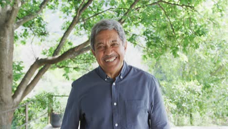 Portrait-of-happy-senior-man-in-blue-shirt-is-standing-against-trees