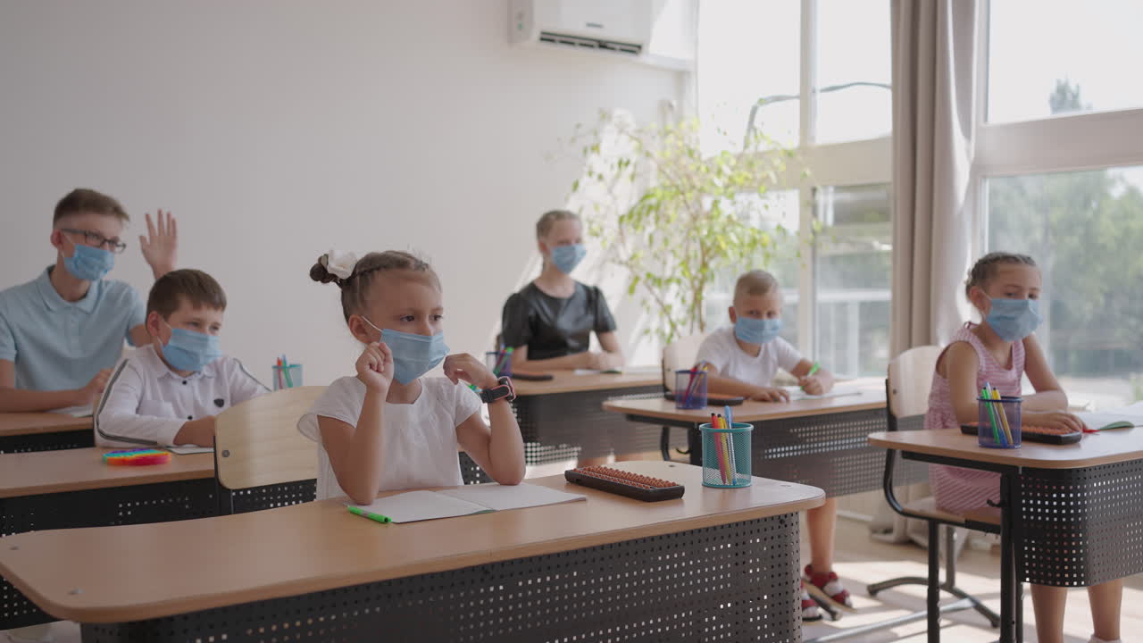Raise your hand to answer the teacher's question. Multi-ethnic group of children with face masks at school during COVID-19 pandemic.