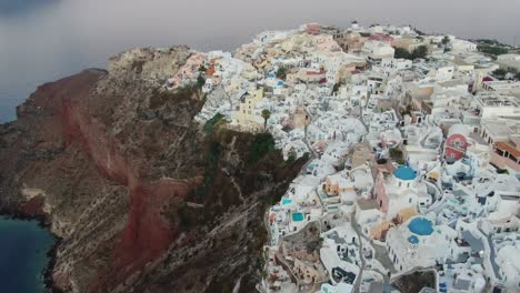 Drone-view-in-Greece-flying-over-Santorini-with-Oia-town-white-houses-and-blue-roofs-on-a-cliff-next-to-the-mediterranean-sea-at-sunrise