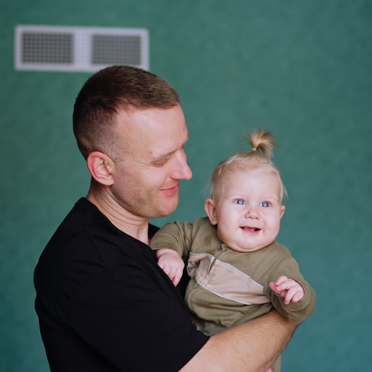 Uomo tiene in braccio un bambino biondo carino. Il grazioso bambino guarda avanti attentamente sorridendo adorabilmente.