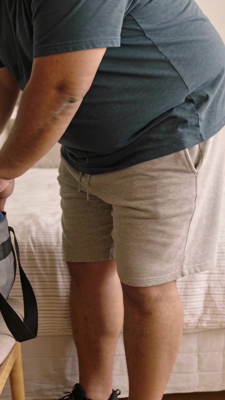 Vertical video: Seeing bag, man in teal shirt tan shorts leaning, packing bag on chair in bedroom
