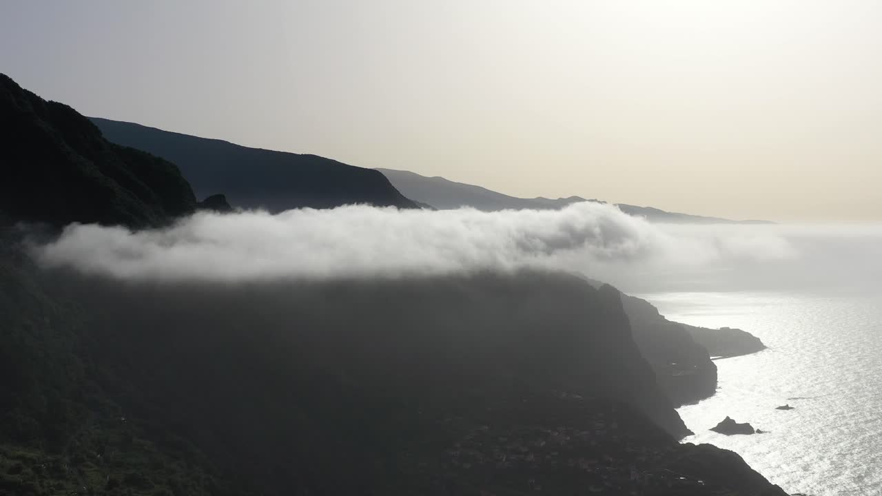 vista aérea de la costa de la isla de madeira, portugal, nubes bajas sobre el océano bajo colinas escarpadas y horizonte brumoso, toma de avión no tripulado