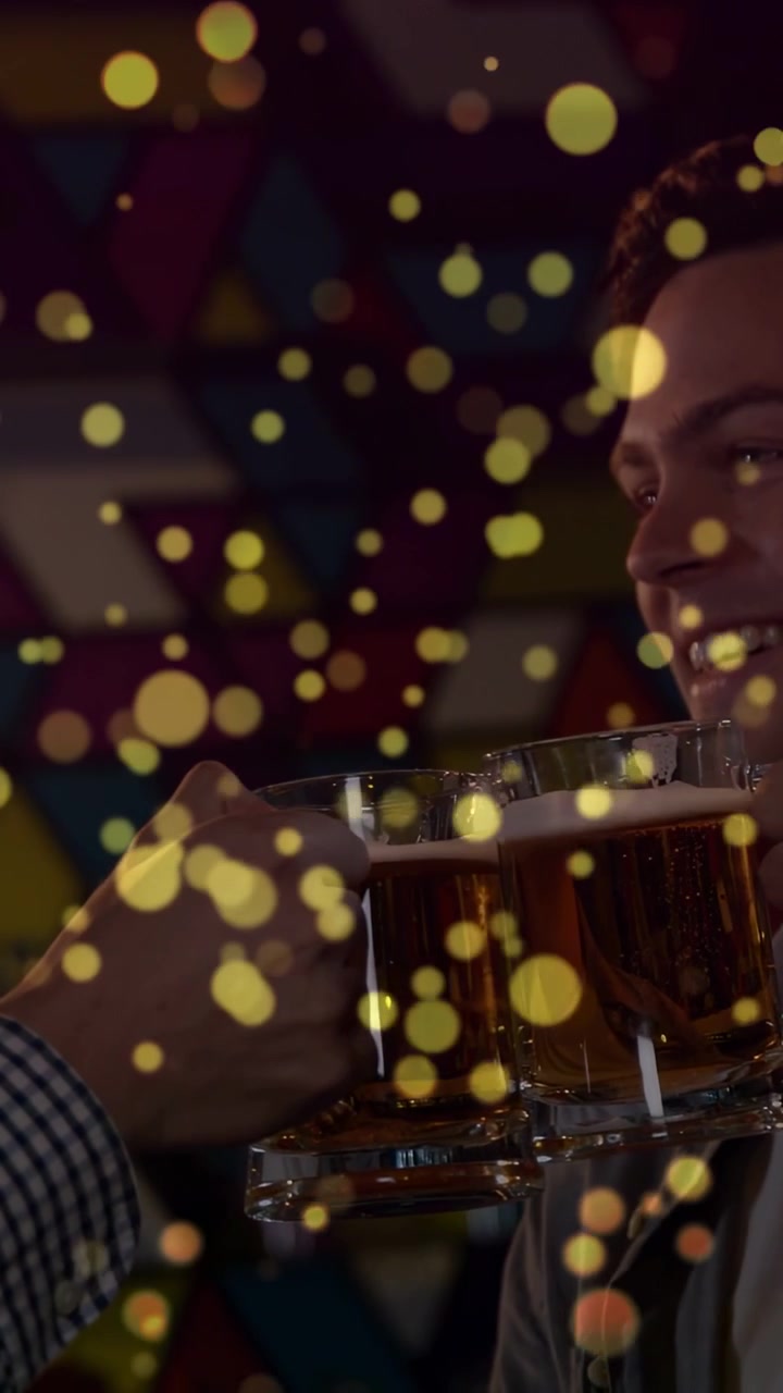 Video verticale: Due uomini che alzano boccali di birra al bar, mani che brindano, boccali che si abbassano sotto un bokeh dorato.