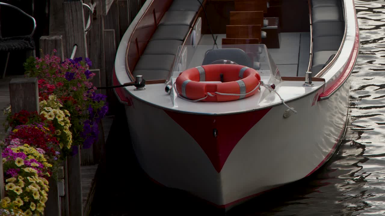 White canal boat leaves flower-lined pier, gentle water ripples, soft natural daylight, smooth camera pan