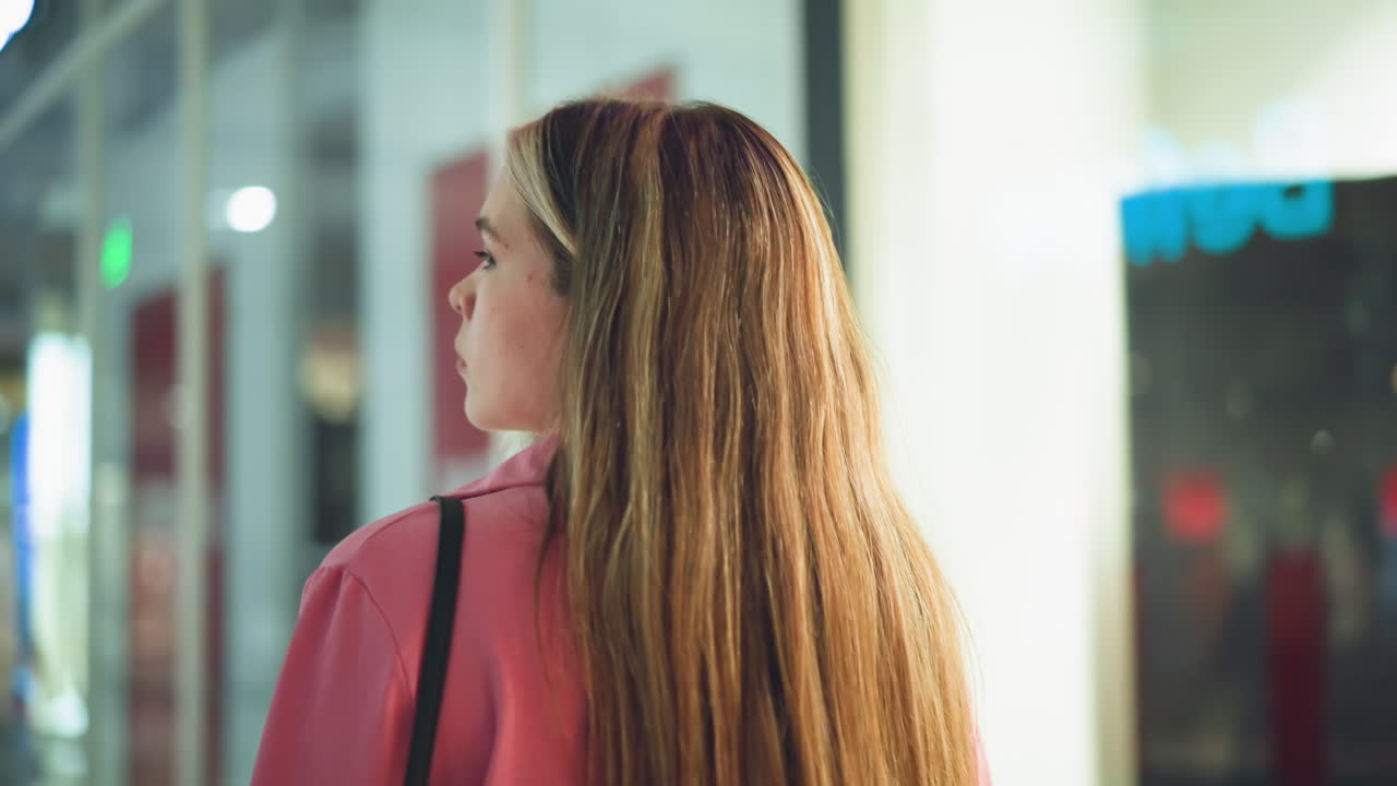 분홍색 드레스를 입은 여자가 쇼핑몰을 걸어가며 어깨를 뒤집어보고, 사람들과 쇼핑몰의 불빛의 반사물이 의 유리 창문에서 보인다.