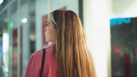 dama de vestido rosa caminando por un centro comercial, mirando hacia atrás por encima de su hombro, los reflejos de la gente y las luces del centro comercial son visibles en las ventanas de vidrio por el lado