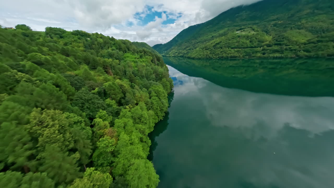 Lush green forest surrounds the serene Lago di Levico, reflecting the cloudy sky, aerial view
