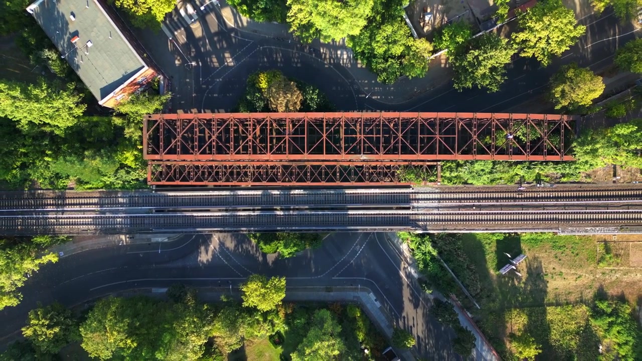 Ponte ferroviario arrugginito che passa sopra i binari del treno in un contesto urbano verde lussureggiante a Berlino, Germania, infrastruttura di trasporto. Spettacolare volo aereo con vista verticale a volo d'uccello da drone.