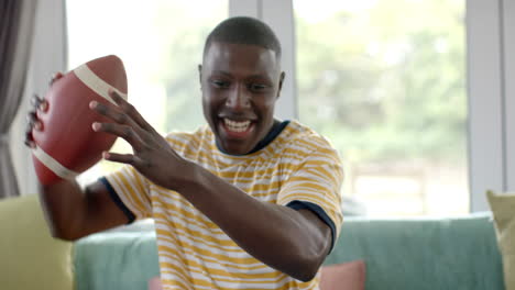 happy african american man on sofa watching rugby match at home, slow motion