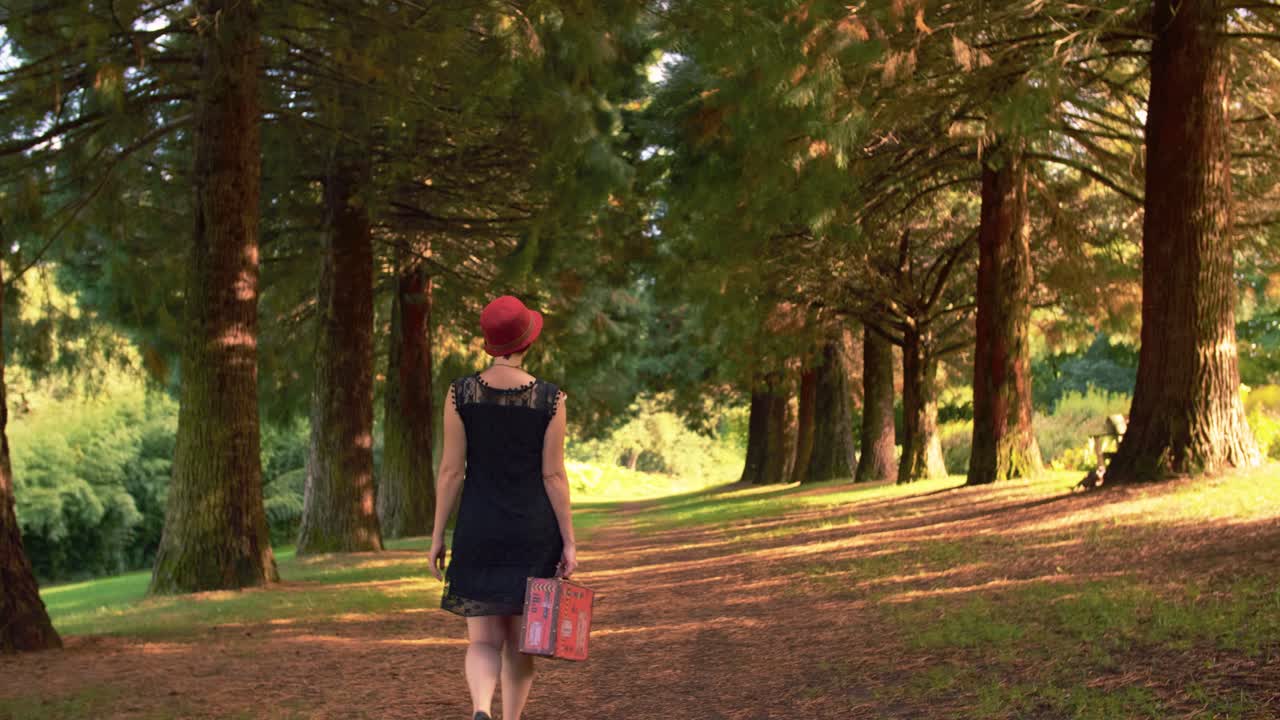 빨간 모자와 검은 드레스를 입은 백인 젊은 여성이 빨간 빈티지 케이스를 손에 들고 카메라 프레임을 걷고 있다