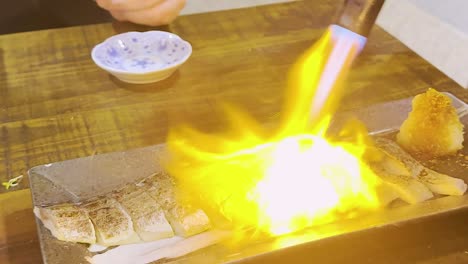 a chef expertly sears fish with a blowtorch, creating a dramatic flame effect on a wooden table.
