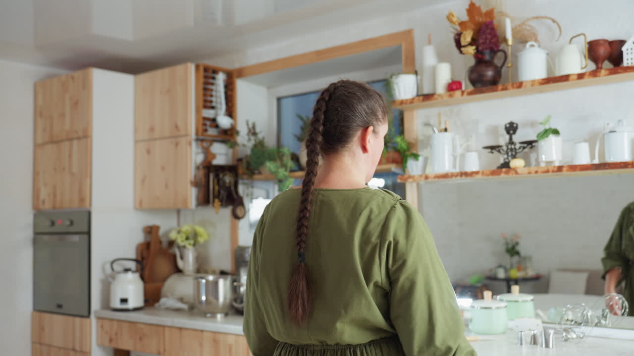 Cook in green gown with long braided hair walks into cozy kitchen filled with wooden cabinets, hanging utensils, potted plants, white teapots, and baking tools arranged neatly on counter and shelves