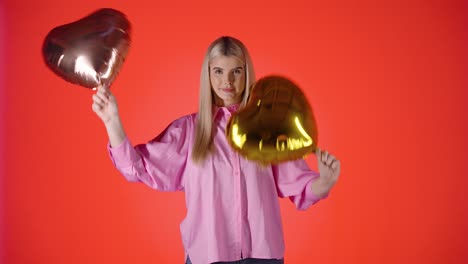 Mujer-Rubia-Sosteniendo-Agitando-Dos-Globos-Dorados-En-Forma-De-Corazón,-Humor-De-San-Valentín-Con-Fondo-De-Color-Rojo,-Foto-De-Estudio