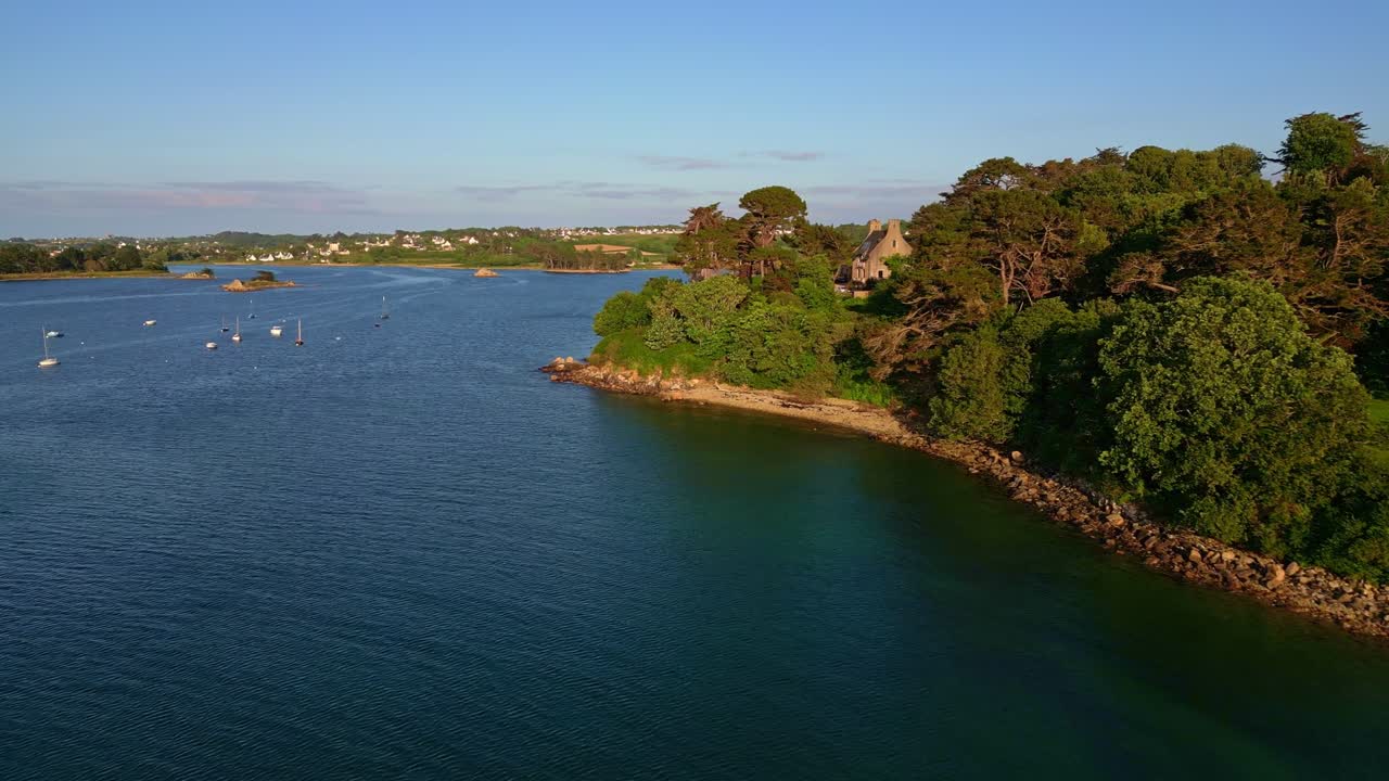 Drone flies over the sea near Port-Blanc, showing trees, shoreline, anchored boats, and an isolated house. Brittany, France