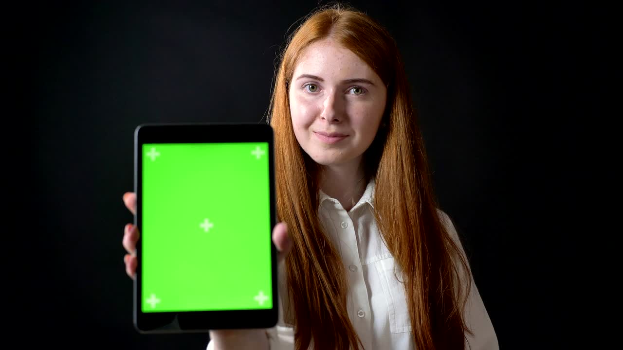 Charming ginger woman showing screen of tablet with charomakey into camera, isolated on black background