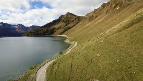 Luftaufnahme-Einer-Bergstraße-An-Einem-See-In-Der-Schweiz-Im-Herbst-Mit-Farbigem-Kiefernwald,-Bewölkter-Herbsttag-Mit-Schneebedeckten-Bergen,-Lago-Ritom