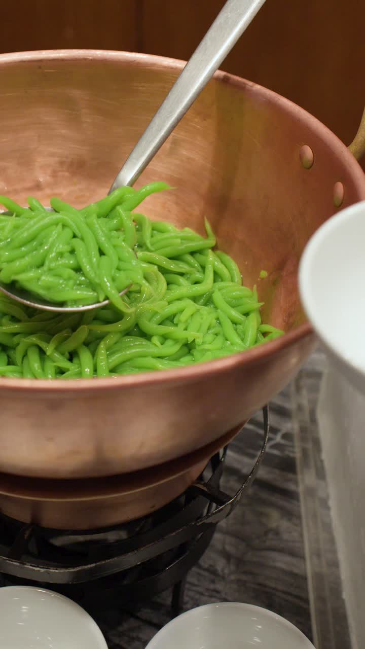 A person scoops green pandan noodles from a copper pot into a white bowl