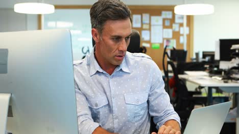 Male-executive-working-on-laptop-and-computer-at-desk