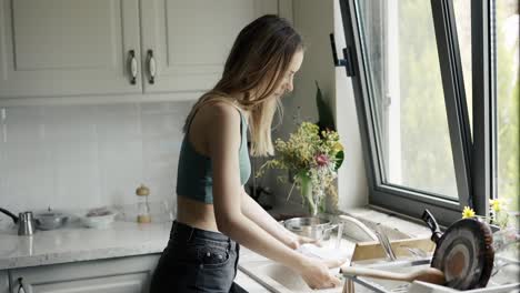 woman washing dishes under running water in sink at bright kitchen