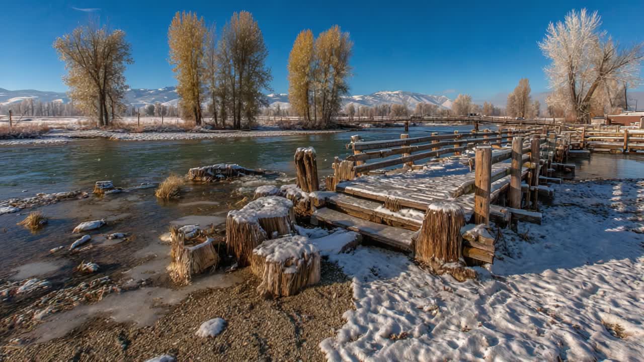 Sereno paesaggio invernale che mostra strutture in legno coperte di brina sulle rive di un fiume calmo, circondato da terreno innevato e alberi maestosi sotto un cielo azzurro limpido