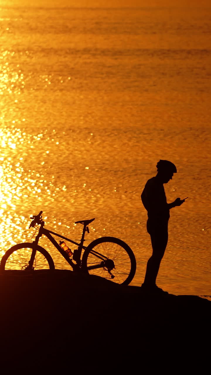 Silhouette of a cyclist and a cat near the river at sunset. Amazing golden path from the setting sun in the water and a man with his bike outdoors. Vertical video