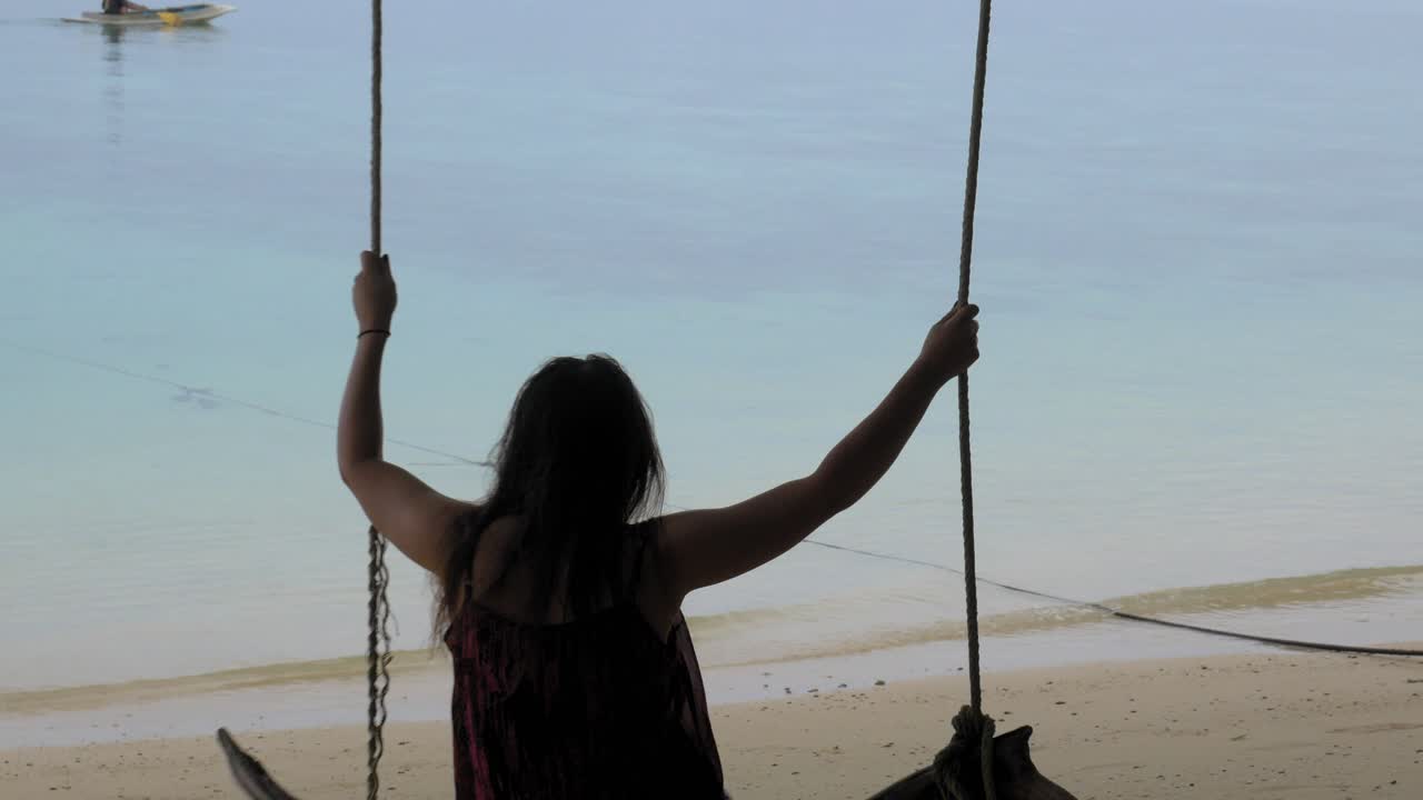 una mujer se sienta y luego se balancea en un columpio de madera mirando un kayak en el mar