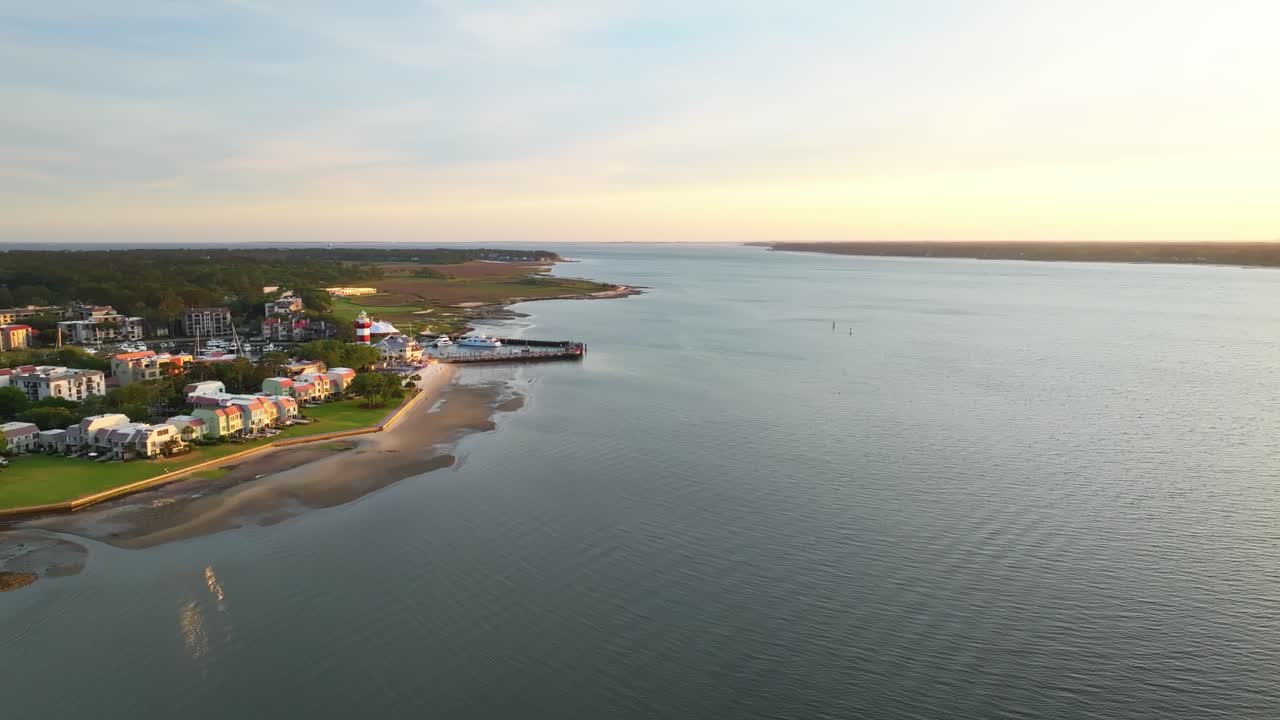 Glide over boats and lighthouse at harbor town pier as sun dips near horizon, panoramic aerial of Harbour Town Pier South Carolina USA
