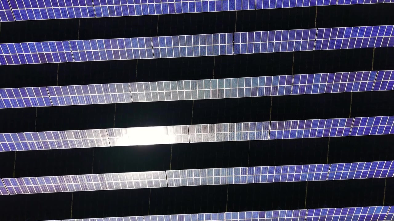 Sun reflection bleaches center panels while surrounding rows show deep blue against dark shadows. Close aerial view of solar panel rows with sun glare reflection, Bulgaria