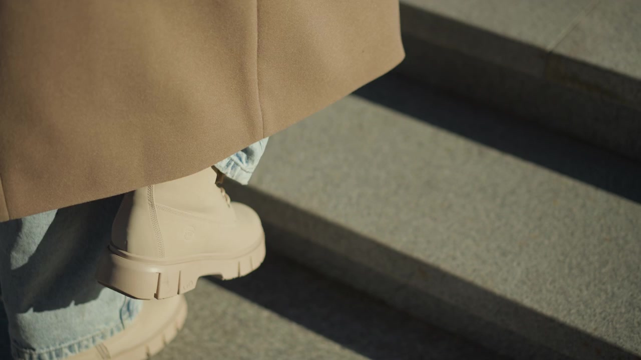 Primer plano de una persona bajando escalones de hormigón con botas beige y vaqueros azul claro, con iluminación suave. Se ven las piernas de la persona llevando vaqueros remangados y un abrigo hasta el tobillo.