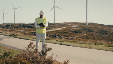 a wide shot of a renewable energy engineer in a reflective vest walking on a field of renewable energy generators, using a tablet to inspect wind turbines, showcasing the role of technology