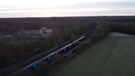 Drohnenaufnahmen-Eines-Zuges,-Der-Den-Bahnhof-Verlässt,-Mit-Einem-Sonnenaufgang-Im-Hintergrund