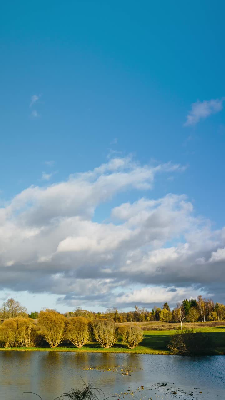 Vertical timelapse video of fast pace cloudscape in autumn. Verdant countryside scenery with pond and forest in the background.