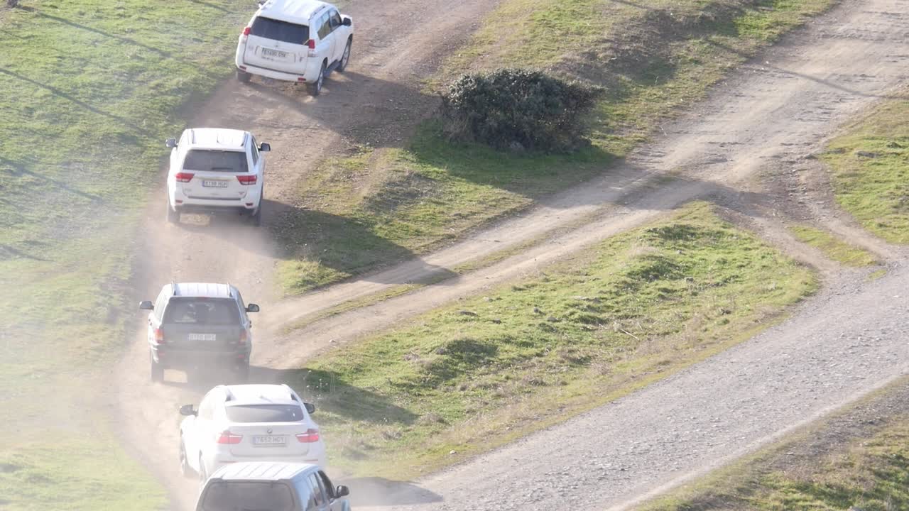 Inquadratura statica dall'alto di diversi SUV che percorrono una strada sterrata tortuosa. Nuvole di polvere e luce mattutina in un paesaggio rurale verdeggiante.