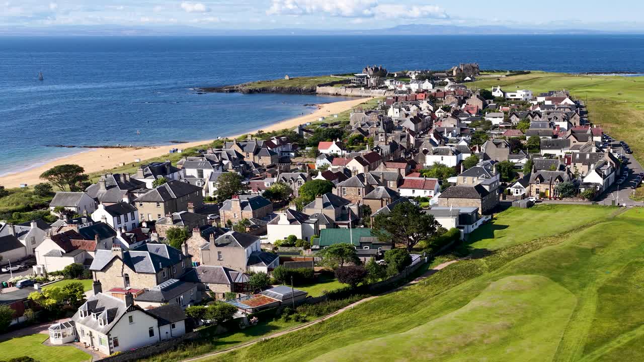 Drone pans above seaside village, green golf course, and coastline under bright daylight, wide perspective
