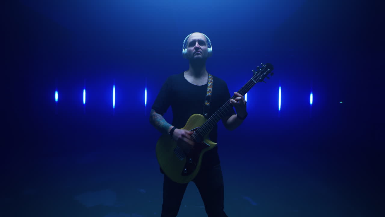 Confident Guitarist Performing against Bright Lamps Tracking Shot of Self Assured Man in Headphones Walking towards Camera and Playing Electric Guitar against Vivid Flickering Lamps in Dark Room