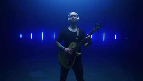confident guitarist performing against bright lamps tracking shot of self assured man in headphones walking towards camera and playing electric guitar against vivid flickering lamps in dark room
