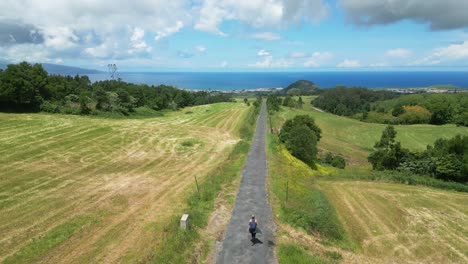 hiker walks along a long paved path through lush green fields towards the distant ocean