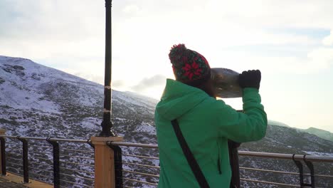 Frau-In-Einer-Grünen-Jacke,-Die-An-Einem-Sonnigen-Tag-An-Einem-Aussichtspunkt-Mit-Einer-Verschneiten-Landschaft-Auf-Ein-Fernglas-Zugeht,-Eine-Reflexion-Der-Sonne-In-Der-Linse