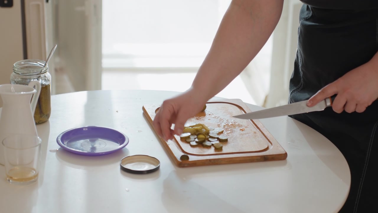 En højrehåndet person skærer syltede agurker med en kniv på et træskærebræt, mens venstre hånd holder ingrediensen fast; hakkede stykker, et glas, låg og køkkenoverfladen skaber en scene med frisk hjemmeforberedelse.
