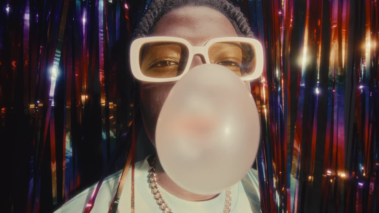 Young Black Man Blowing Bubblegum at Camera against Shiny Tinsel Curtain