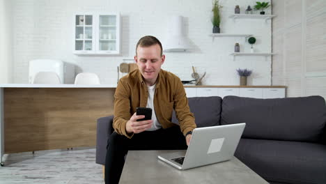 male freelancer working at home. young man sitting on a sofa in front of a laptop and looking into his mobile phone on kitchen background.