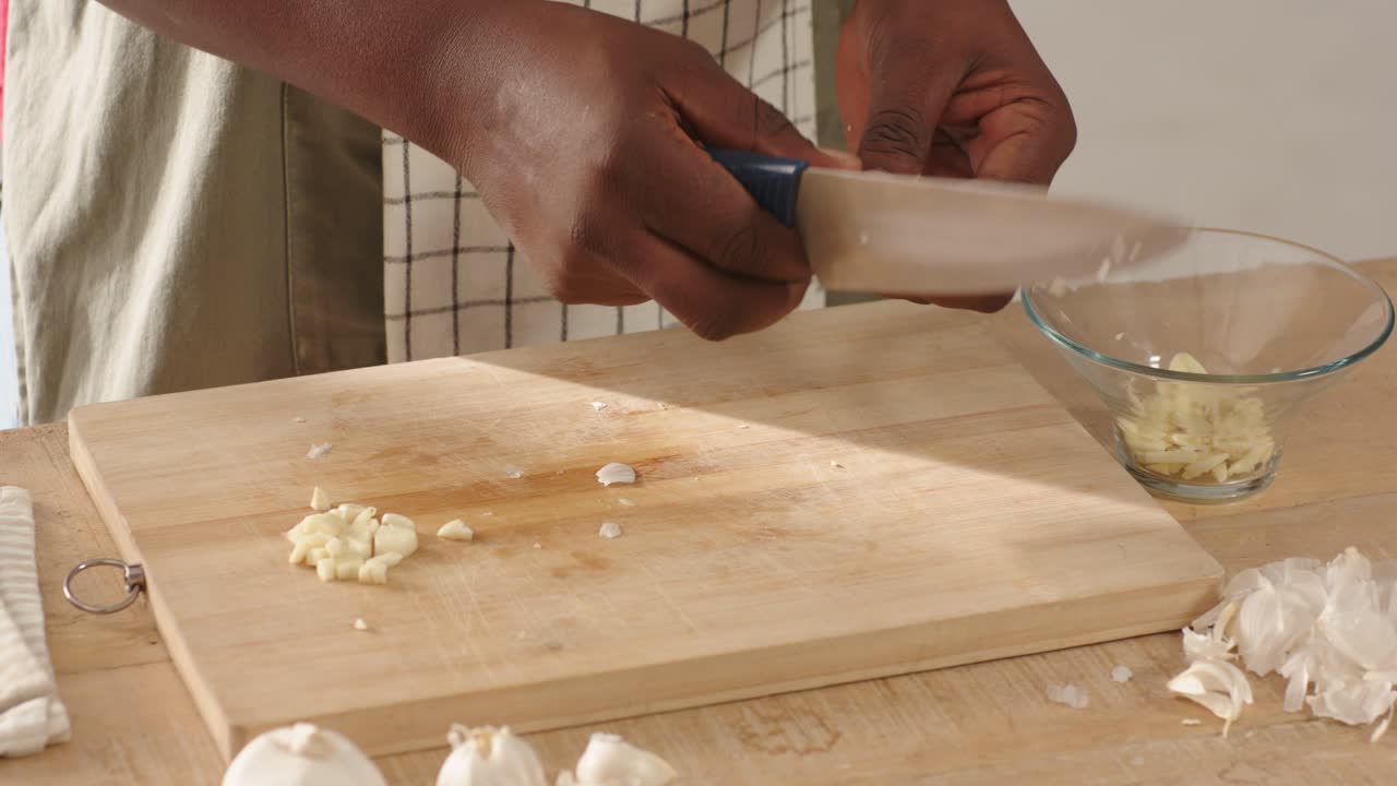 Uomo afroamericano che sbuccia, affetta e trita l'aglio per cucinare su un tagliere, con un coltello blu e un grembiule.