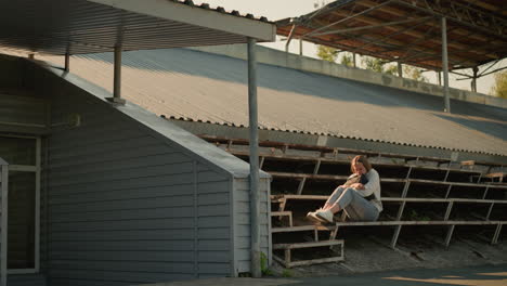 woman seated alone on empty stadium bleachers in a thoughtful pose, hands holding her bag close, metal roof and surrounding greenery provide serene natural background